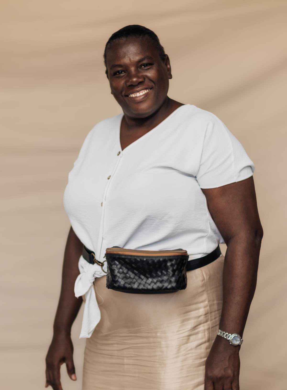 A smiling woman wearing a white short-sleeve button-front top tied at the waist, a beige skirt, and a black woven belt bag, standing against a warm beige backdrop.