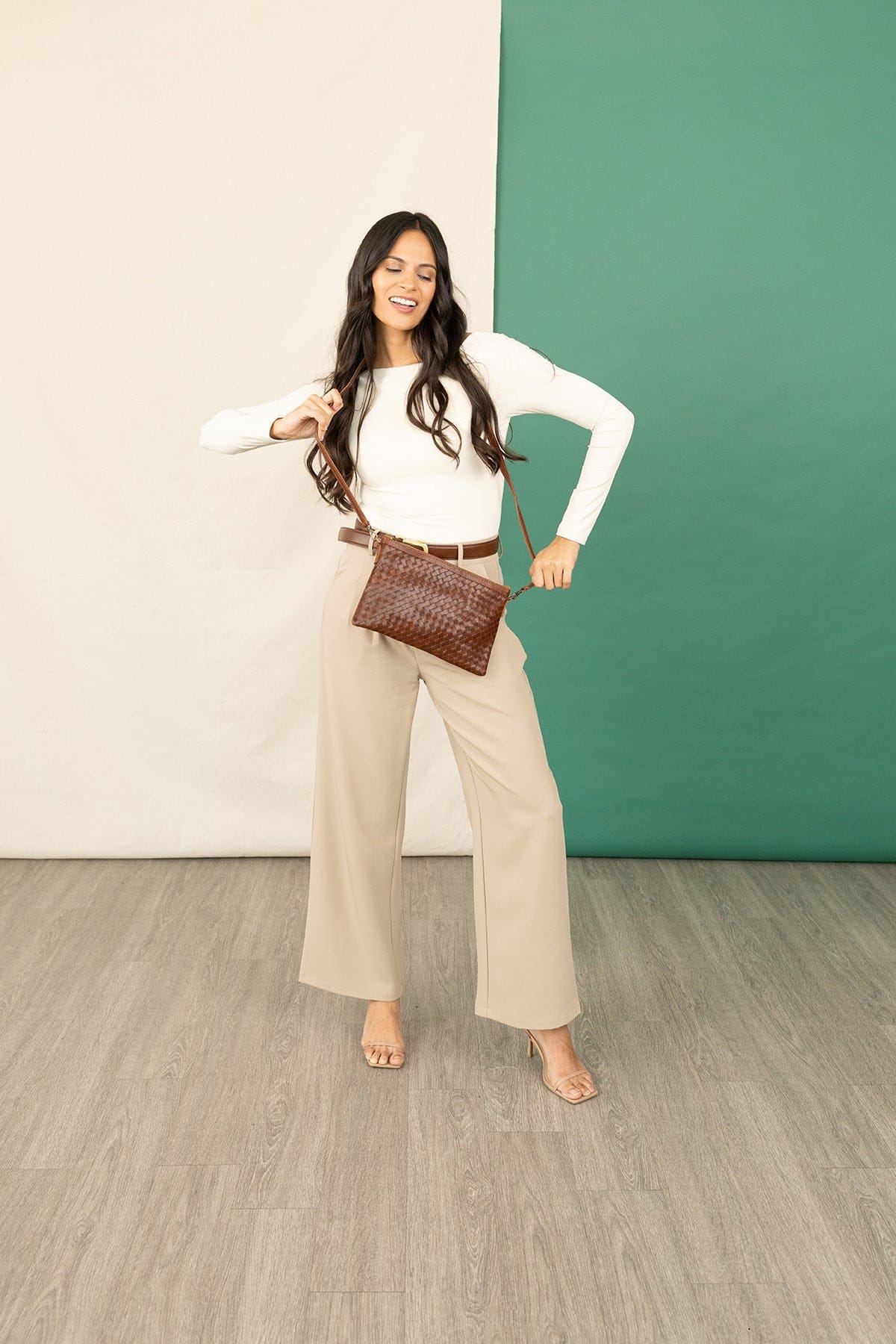 Woman in a white top and beige pants holding a brown woven handbag against a green and beige wall.